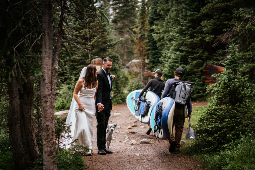 Bride and groom in wedding clothes walk past two people carriyng standup paddleboards at Brainard Lake, Colorado adventure wedding