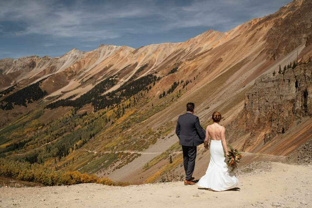 Bride and groom walk down road holding hands with fall colors in background at Telluride elopement.