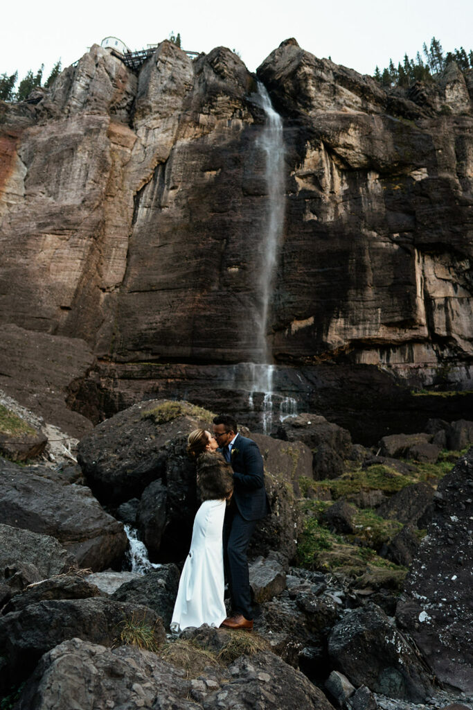 Bride and groom snuggle at base of waterfall in Telluride, colorado at Bridal Veil Falls.