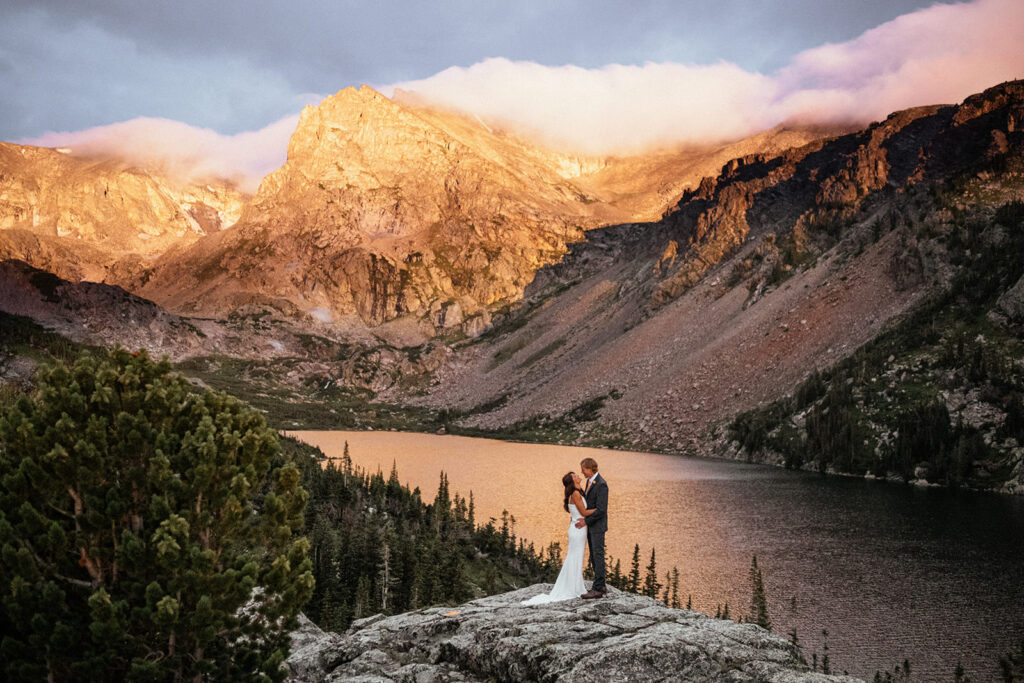 Bride and groom in wedding clothes standing on rock above lake with mountains in the background at Colorado mountain elopement at Lake Isabelle in the Indian Peaks wilderness