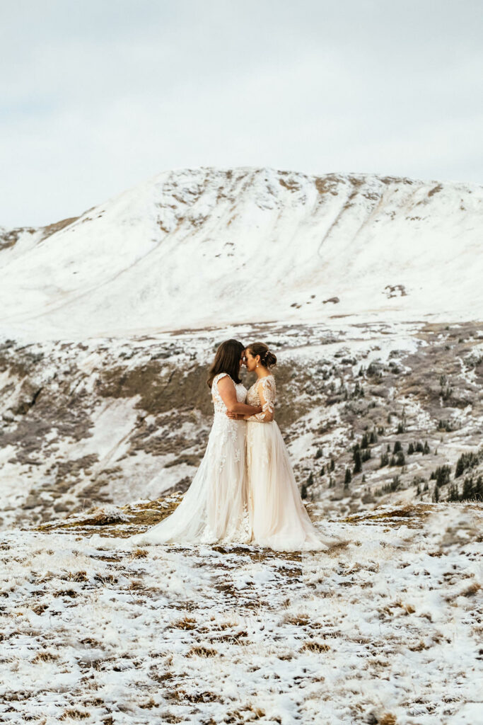 Two brides in wedding dresses snuggle in the snow on top of mountain at Colorado adventure elopement