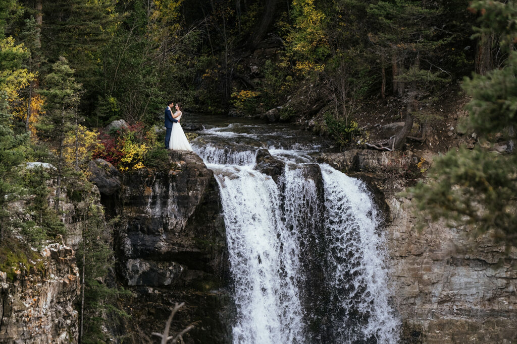 Bride and groom in wedding clothes stand on top of giant waterfall at Colorado elopement in the mountains