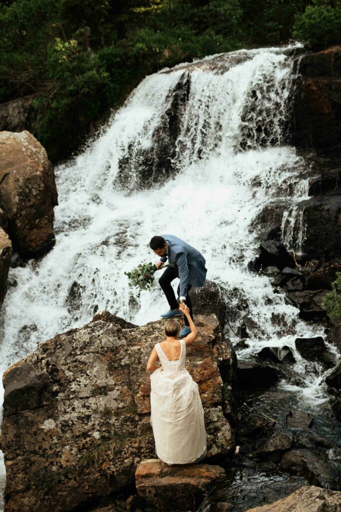 Bride and groom climb onto boulder in the middle of a waterfall at breckenridge colorado elopement