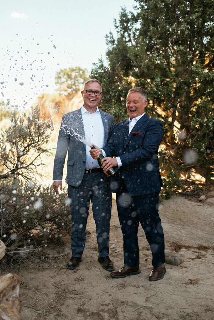 Two grooms in suits pop a bottle of champagne at Colorado elopement